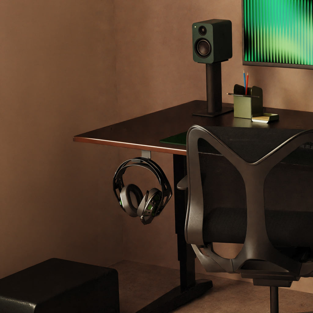 A modern desk setup in a warm brown room, featuring a black desk with a hook holding over-ear headphones, a black ergonomic chair, a speaker, and a computer monitor.