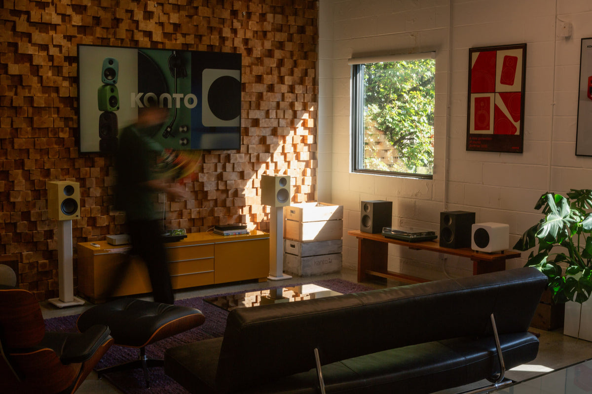 A modern living room scene with a person in motion blur in front of a wall-mounted TV, set against a brown geometric wood-paneled wall with floor-standing speakers and a yellow media console.