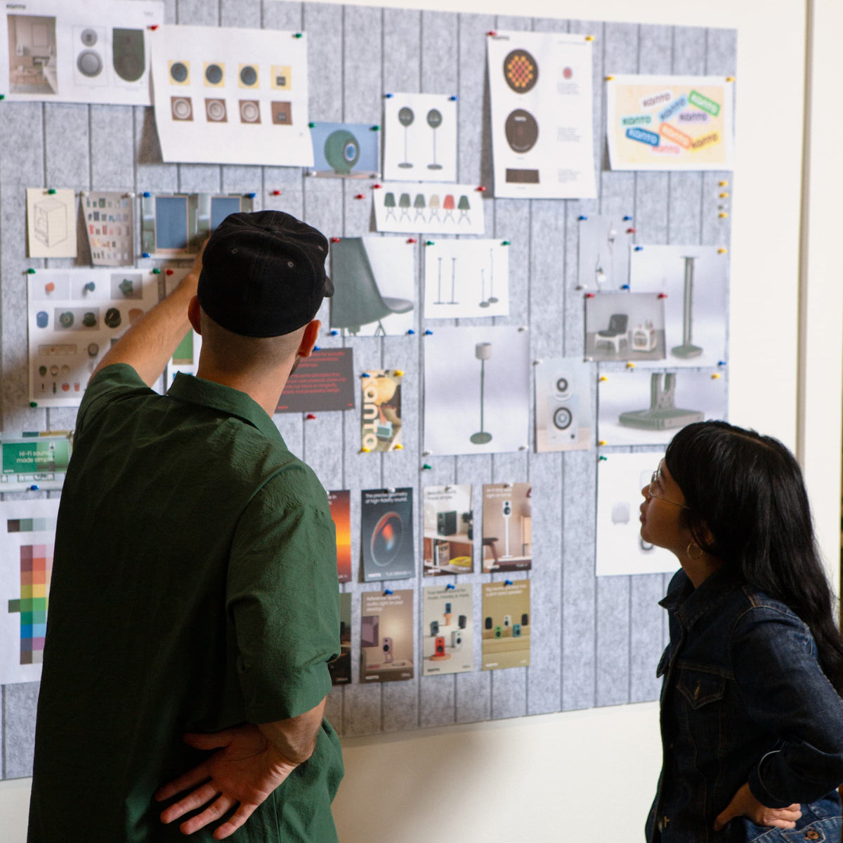 A man in a green shirt and black cap points at a large corkboard filled with design sketches and product photos, while a woman in a denim jacket looks on.