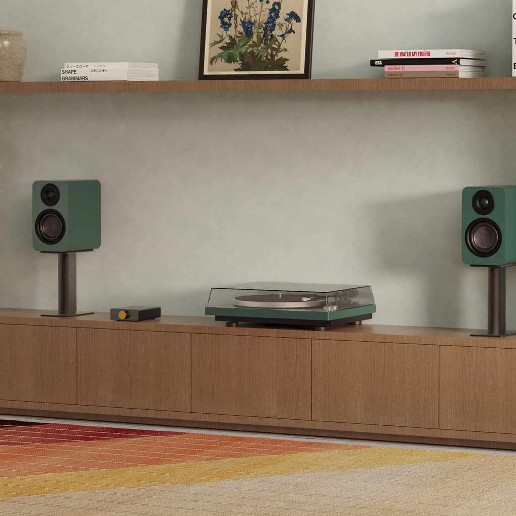 A Obi3 Turntable with a clear dust cover and green accents sits on a wooden media console, flanked by matching green speakers on a minimalist living room setup.