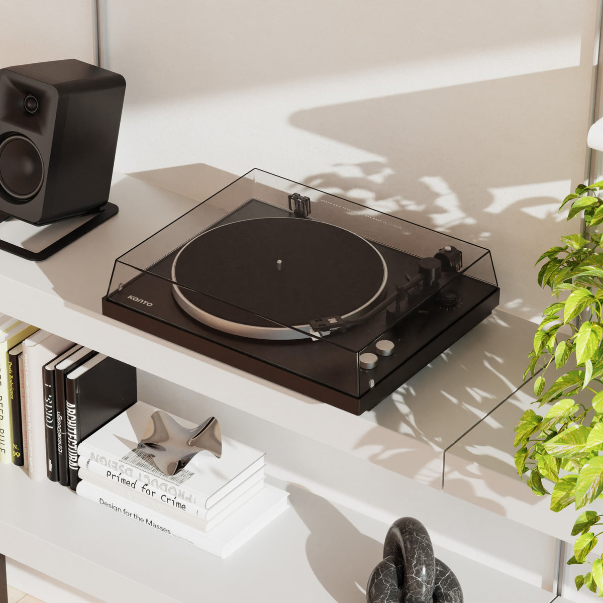 A black Obi3 turntable with a clear dust cover sits on a white shelf beside a black speaker, books, and a green plant in a bright interior setting.