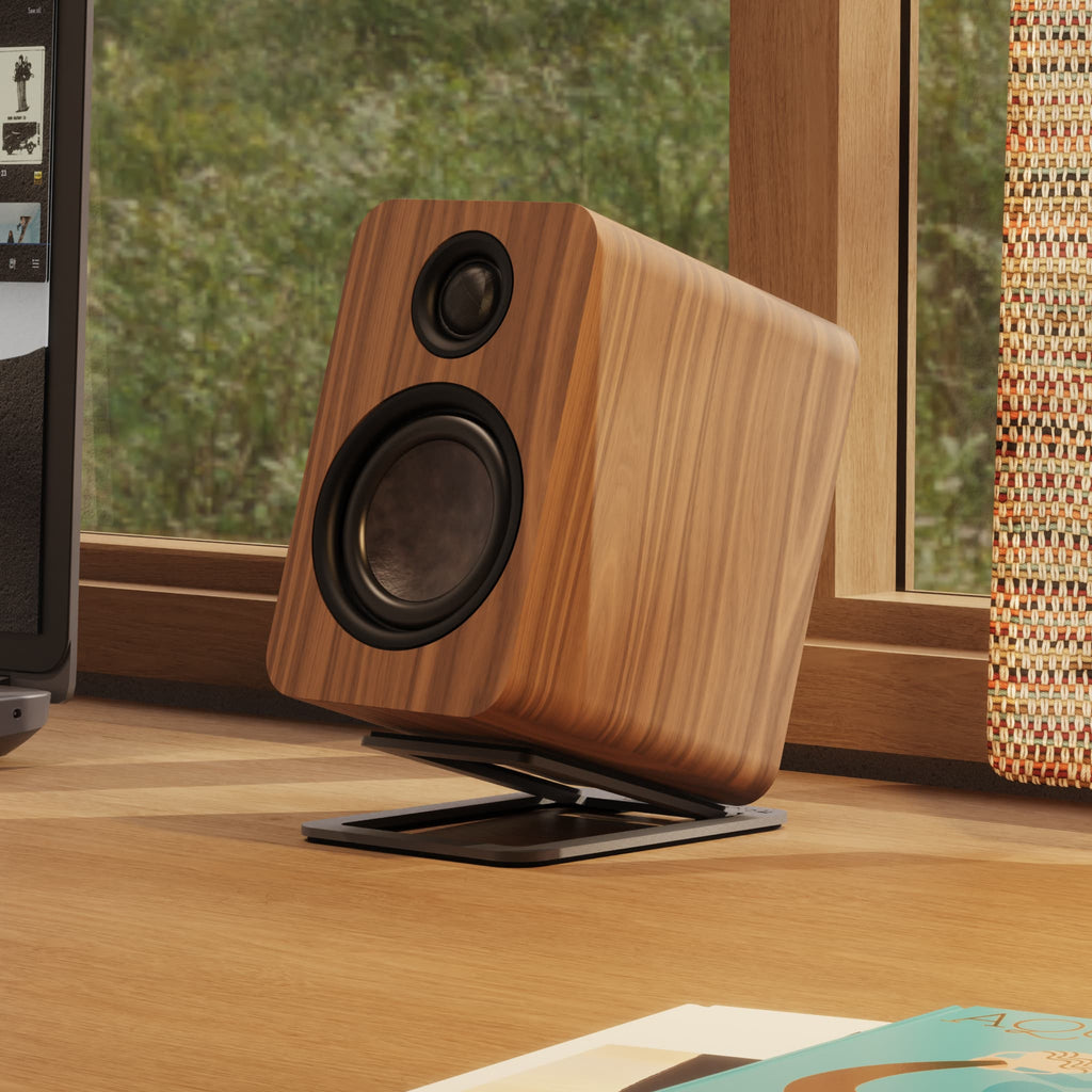 A wooden speaker with a black grille and stand, displayed on a wooden desk in front of a window.