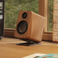 A wooden speaker with a black grille and stand, displayed on a wooden desk in front of a window.