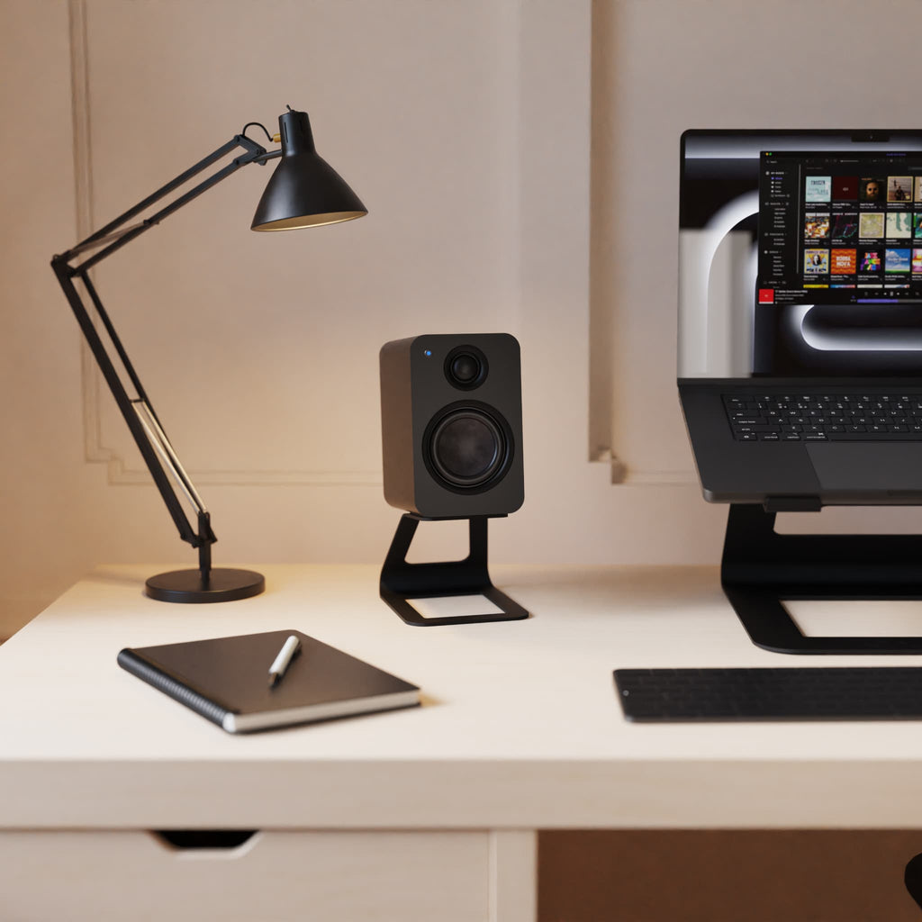 A black speaker on a black stand displayed on a white desk next to a black adjustable desk lamp, a closed notebook with a pen, and a laptop on a monitor stand.