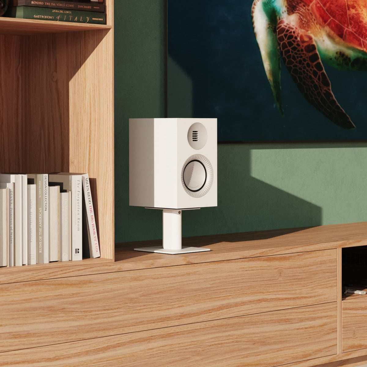 A white Kanto Tuk speaker on a matching white stand, placed on a wooden console in a styled room with a bookshelf, green wall, and TV.