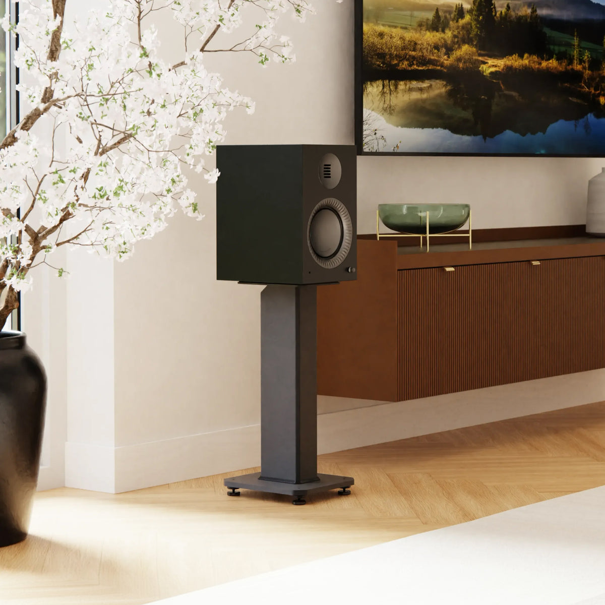 A black bookshelf speaker on a matching black floor stand, set in a modern living room next to a wooden cabinet and a TV.