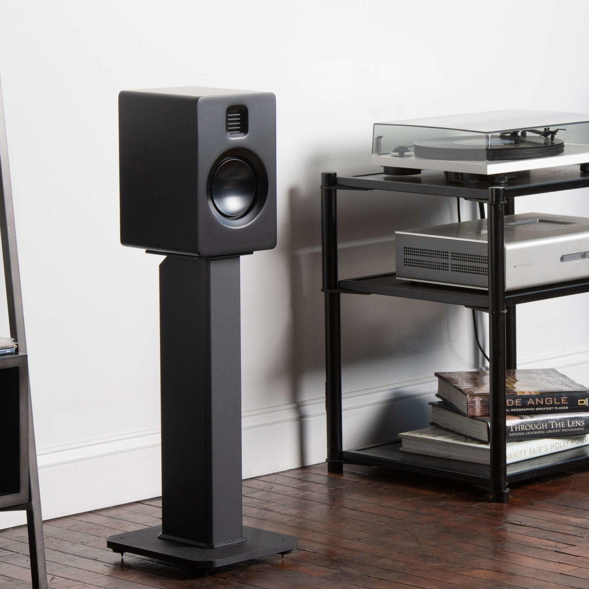 A black studio speaker on a matching SX26 floor stand, shown in a home theater setting next to a black shelving unit with a turntable and audio equipment.