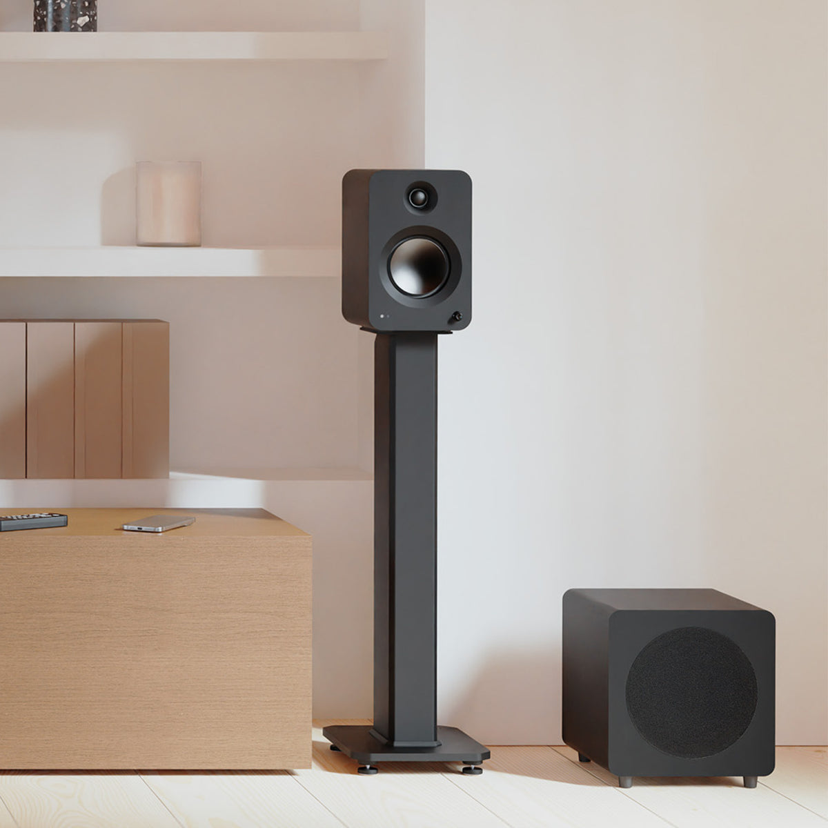 A black bookshelf speaker on a matching floor stand is shown in a minimalist room setting next to a matching floor subwoofer.