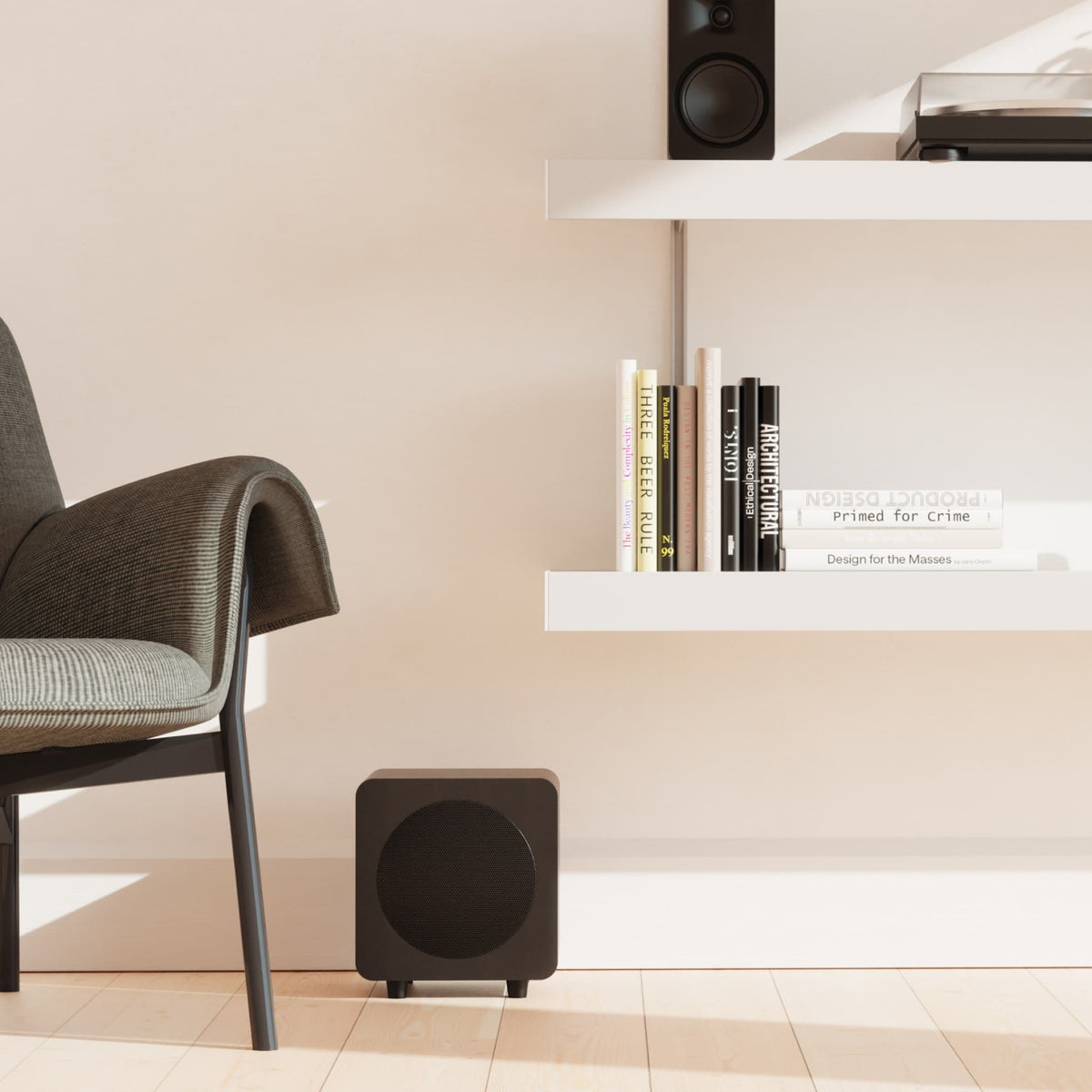 A balck subwoofer sits on a light wood floor in a minimalist room setting, next to a gray upholstered chair and beneath white floating shelves holding books and another speaker.