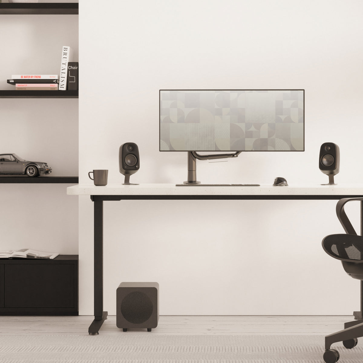 A minimalist home office setup featuring a black standing desk with a wide monitor, two black Uki speakers, a mug, and a subwoofer, set against a clean white wall with built-in shelves on the left.