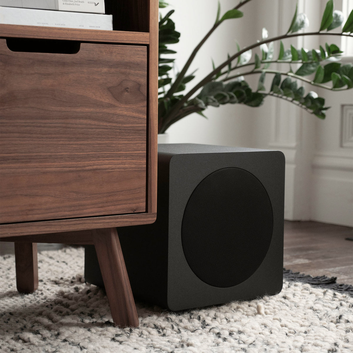 A black square subwoofer speaker with a circular front grille, positioned on a white shag rug next to a wooden nightstand in a modern room setting.