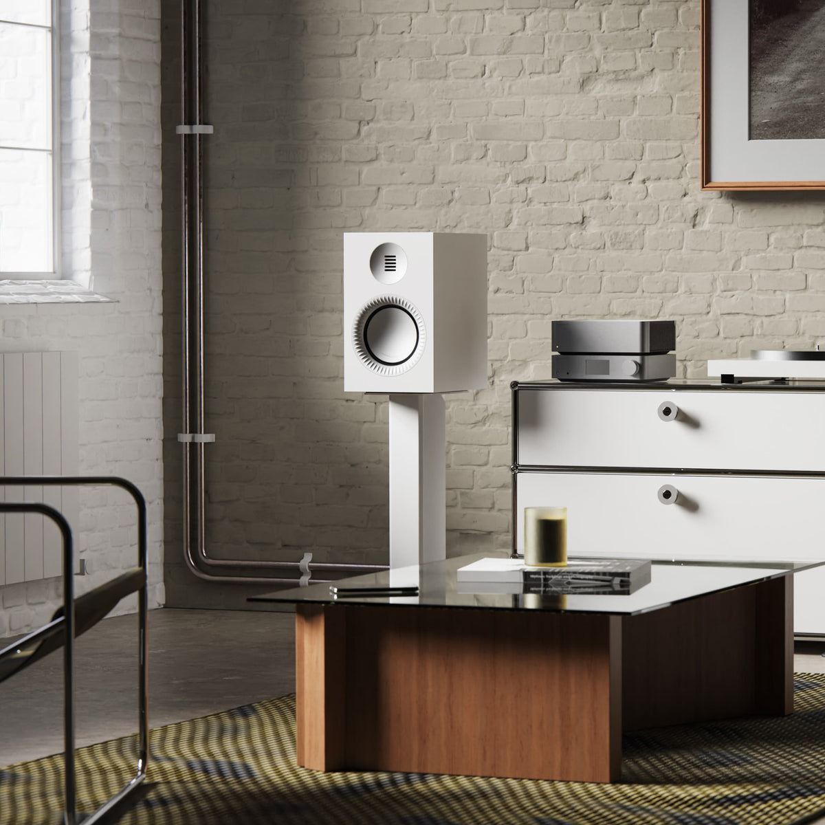 A white Tuk Grand Passive speaker on a matching white display stand in a modern living room setting with a brick wall, wooden coffee table, and white cabinet.