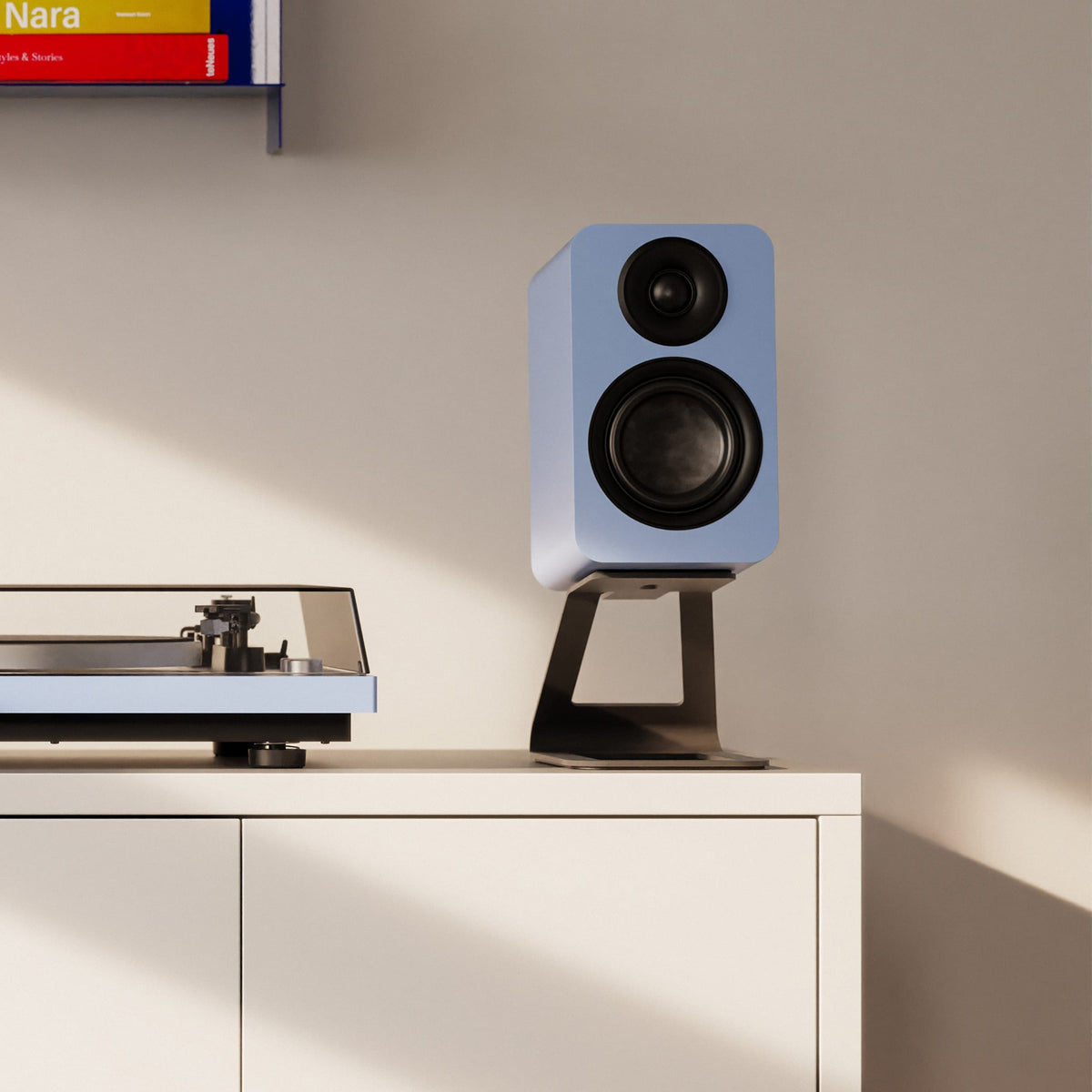 A light blue bookshelf speaker with black drivers on a black stand, displayed on a white cabinet in a minimalist room next to a turntable.