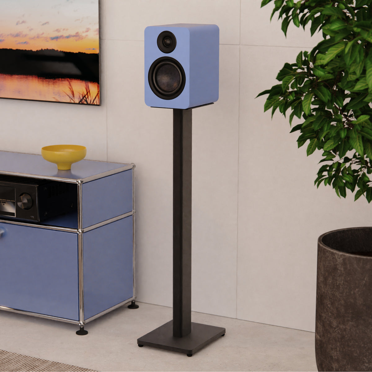 A light blue Yu6 Passive bookshelf speaker mounted on a tall black floor stand, shown in a modern living room setting next to a blue cabinet and a potted plant.