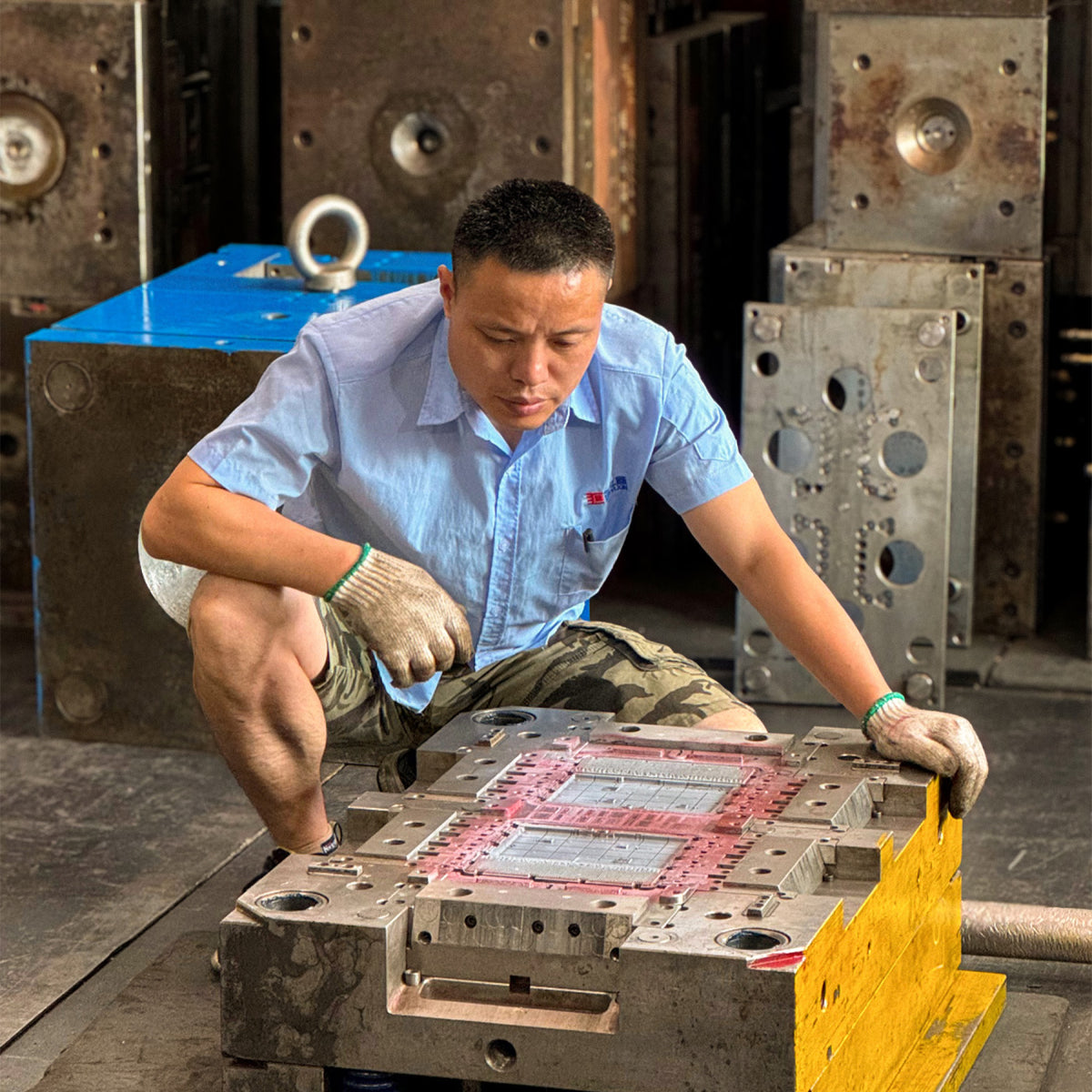 A male worker in a light blue shirt kneels down in a workshop, adjusting a large metal mold. Heavy industrial machinery and metal blocks are visible in the background.