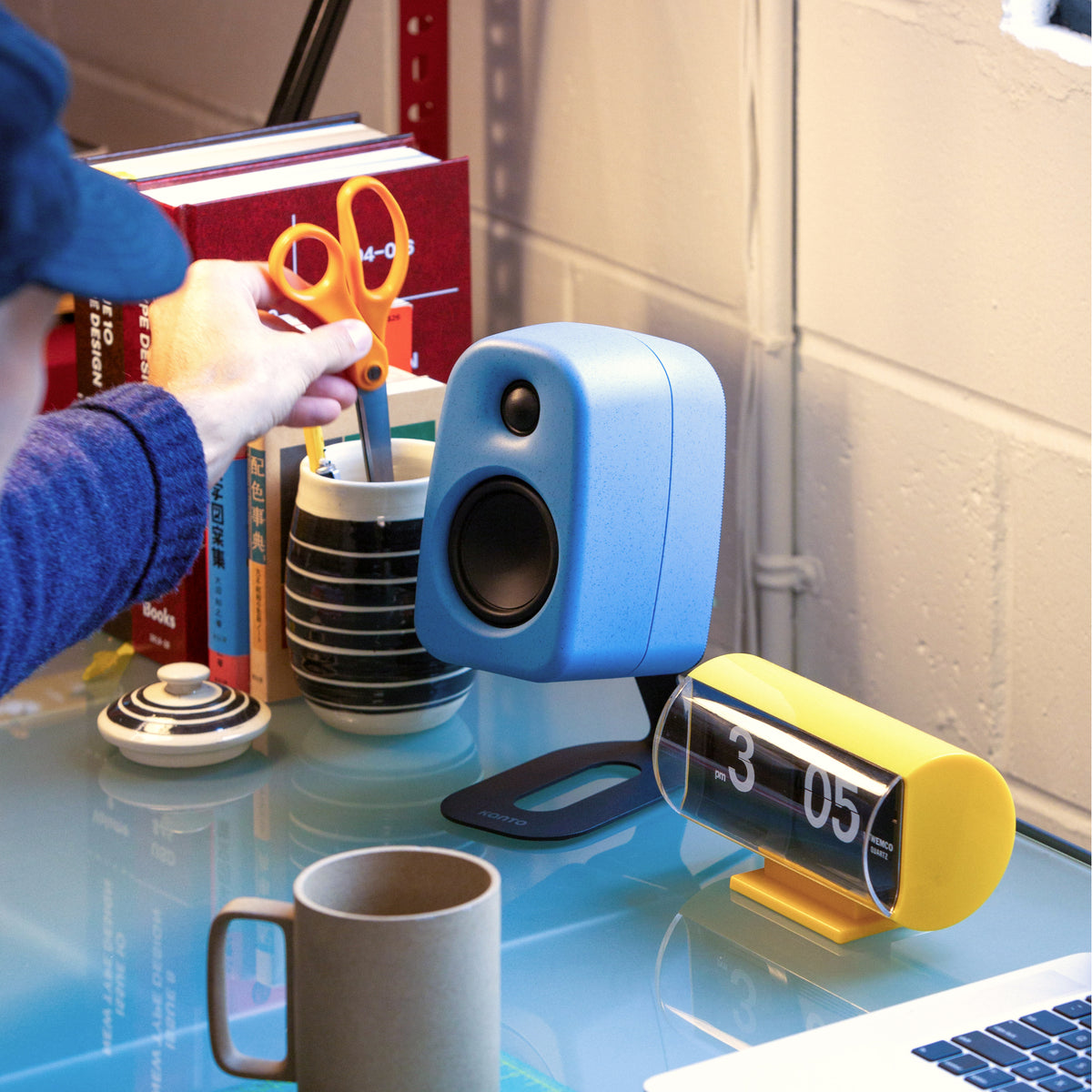 A blue Uki desktop speaker and a yellow desk clock on a glass table in a workspace setting, with a person's hand holding orange-handled scissors in the background.